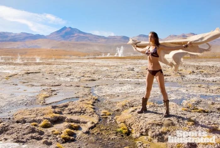 Irina Shayk stands in the desert wearing a brown bikini, brown leather boots and holding a tan blanket up behind her.